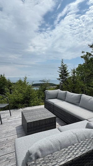 A deck with a grey sofa and a woven ottoman, overlooking a forested area and a distant island.