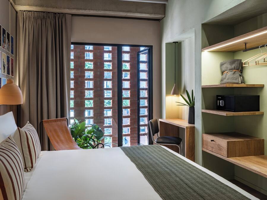Ein Hotelzimmer mit einem Bett, einem Schreibtisch, einem Stuhl, einer Pflanze und Blick auf ein Backsteingebäude durch ein Fenster.