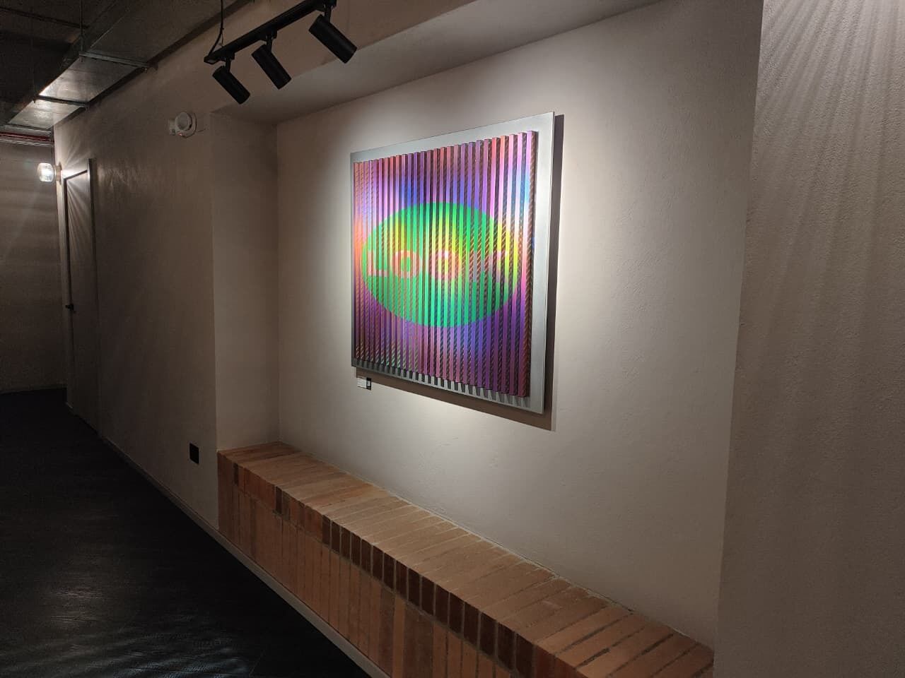 Un couloir avec un banc, une œuvre d’art accrochée au mur et un luminaire au plafond.