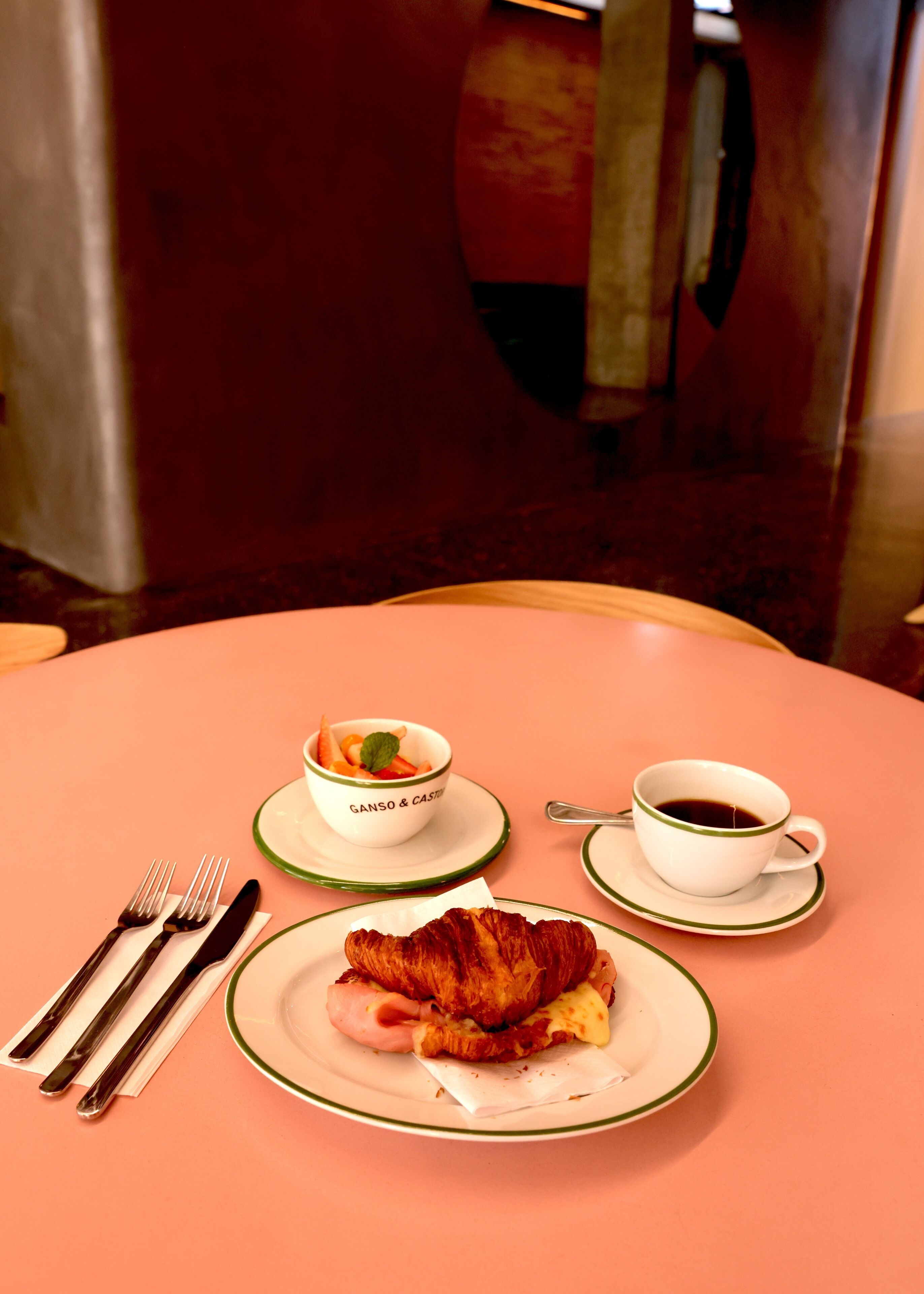 A table with a croissant, a cup of coffee, a bowl of fruit, and cutlery.