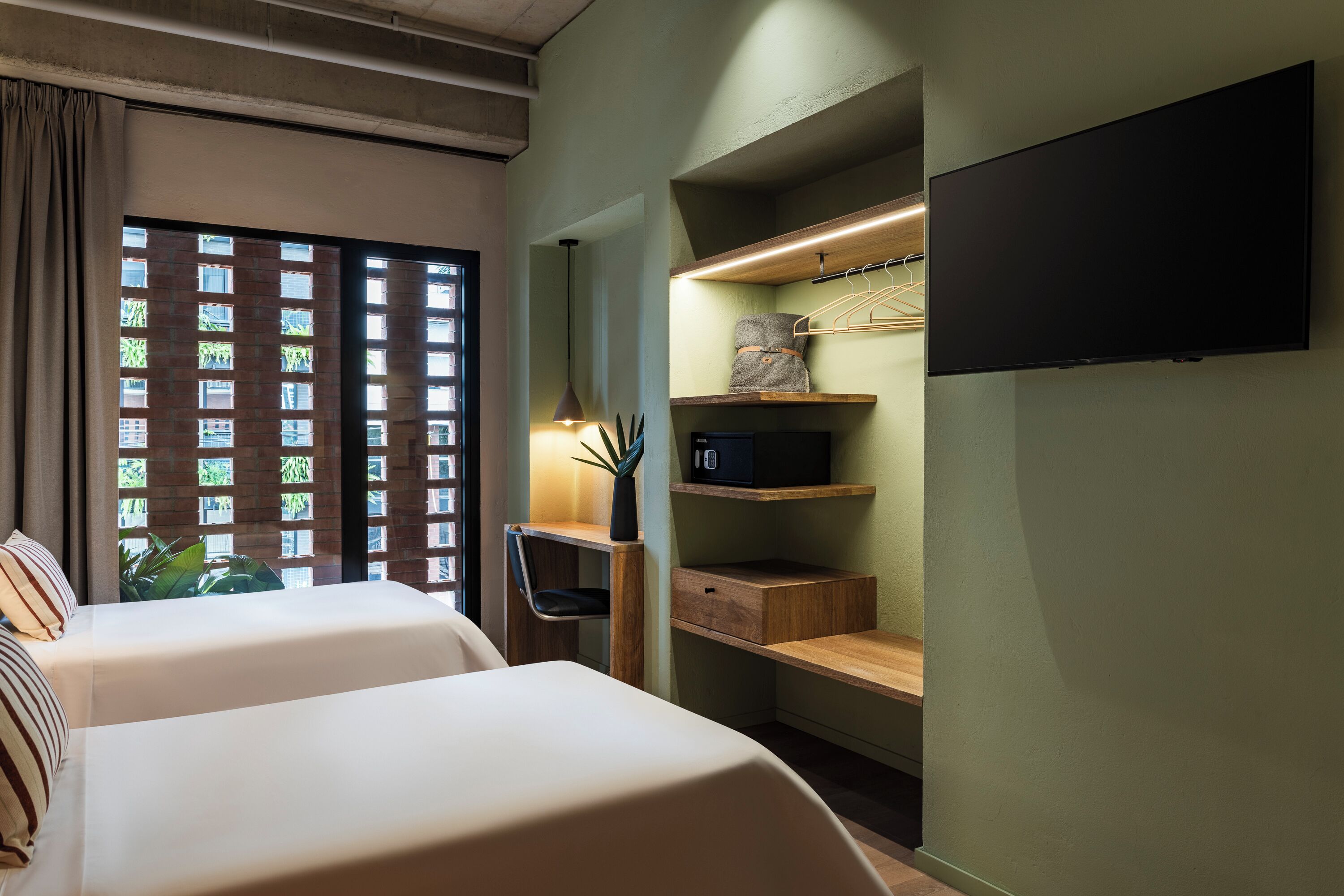 A hotel room with two beds, a desk, a TV, and a window with a view of greenery.