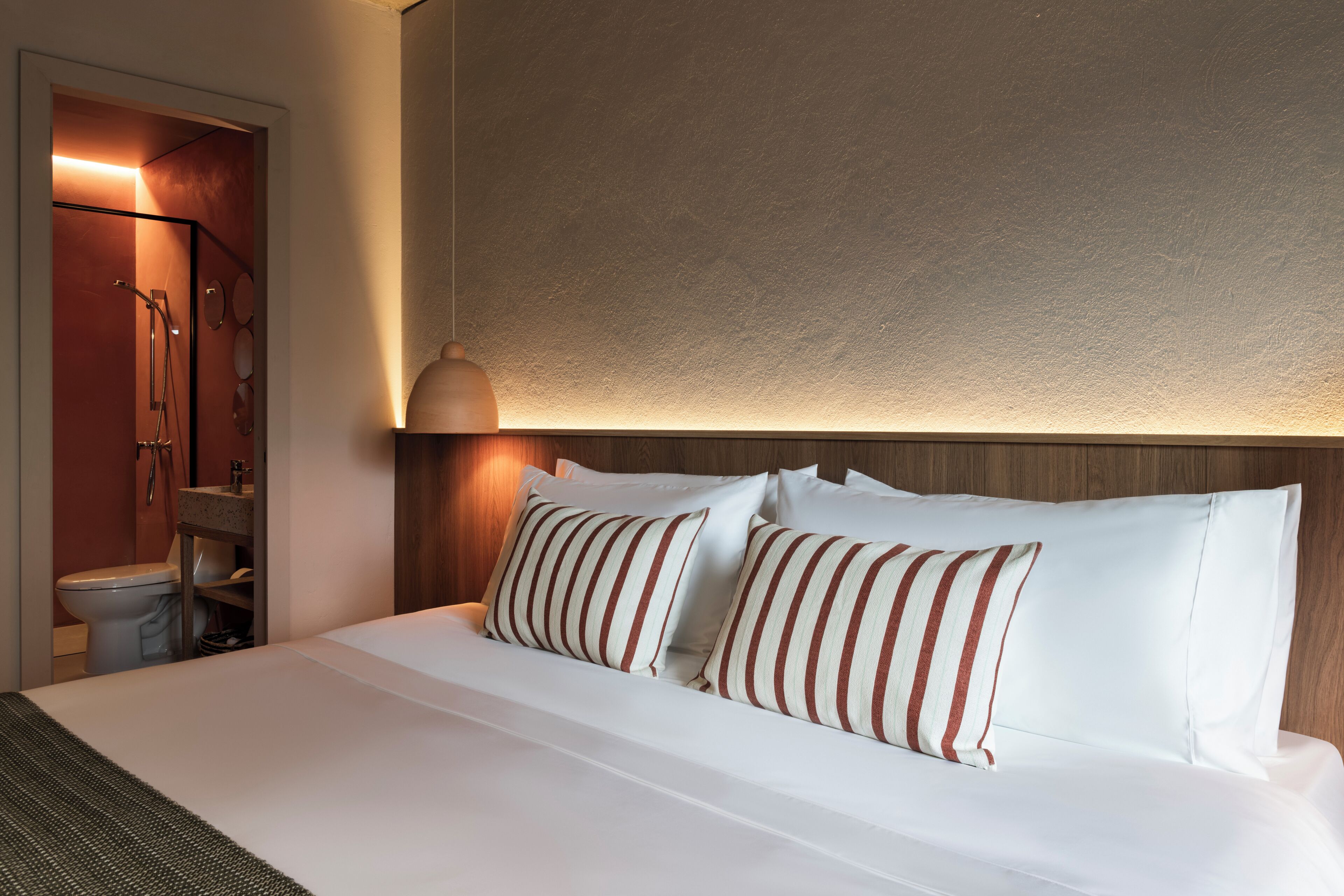 A hotel room with a bed, pillows, a nightstand, a lamp, and a bathroom visible through an open door.
