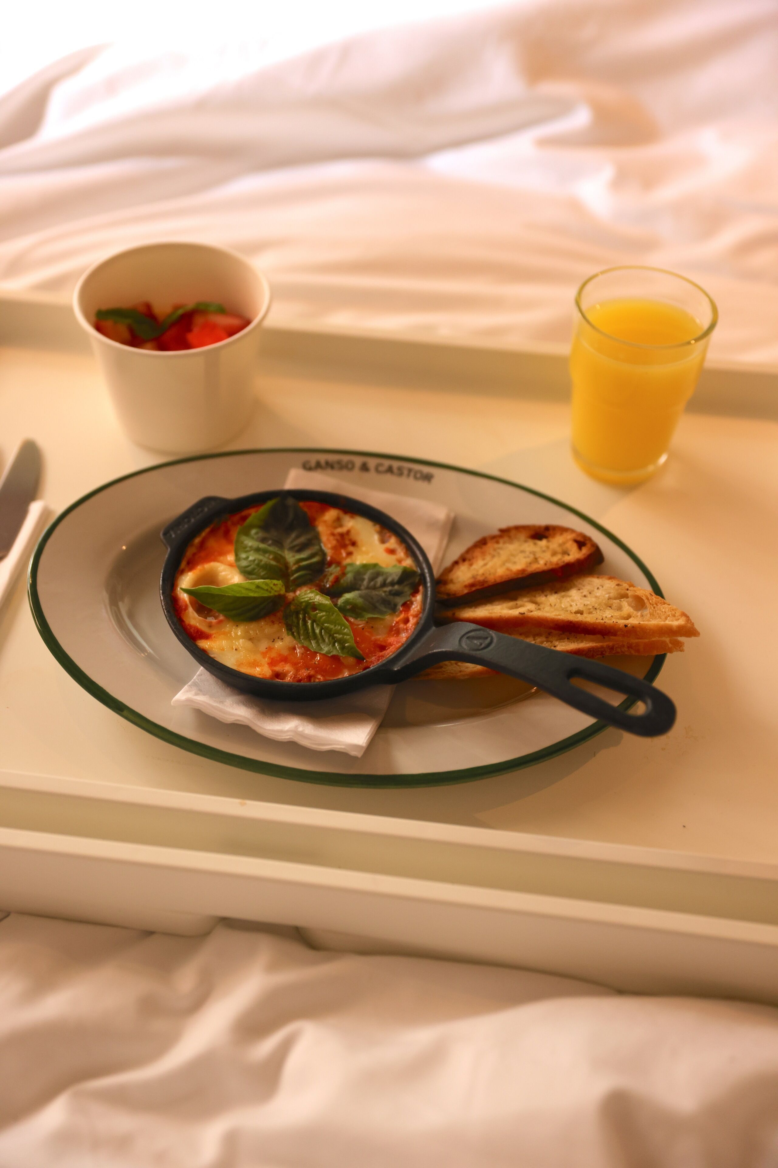 Un plateau de petit-déjeuner avec une poêle remplie de nourriture, un verre de jus d’orange et une tasse de café, posé sur un lit.