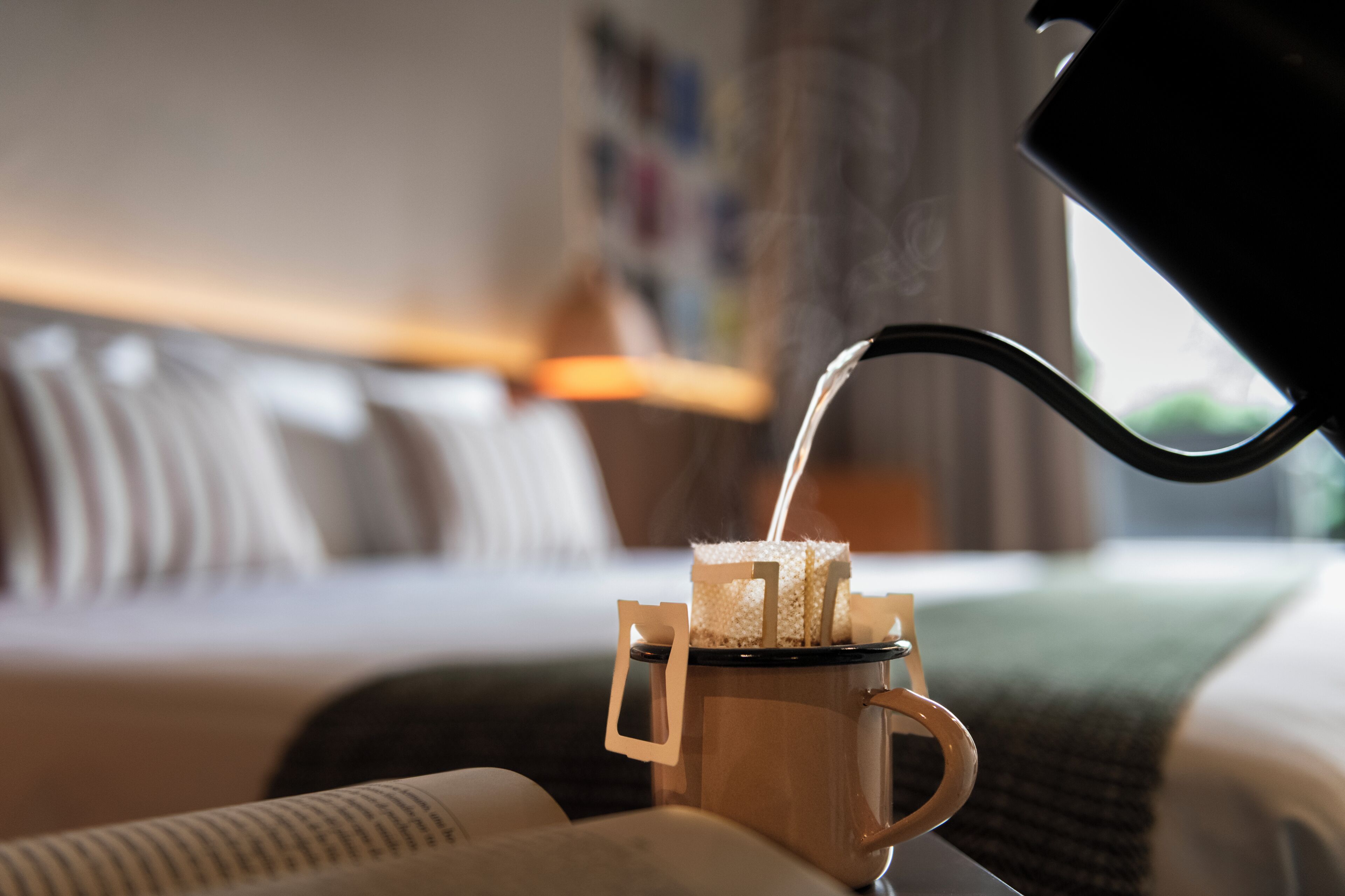 En kaffekanna häller kaffe i en mugg som står på ett hotellbord, bredvid kuddar och en filt.