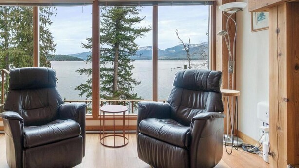 Ein Zimmer mit zwei Ledersesseln, einem kleinen runden Tisch und Blick auf einen See und Berge durch große Fenster.