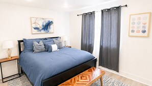 Quarto com cama, mesas de cabeceira, um banco, tapete e uma parede com uma moldura.