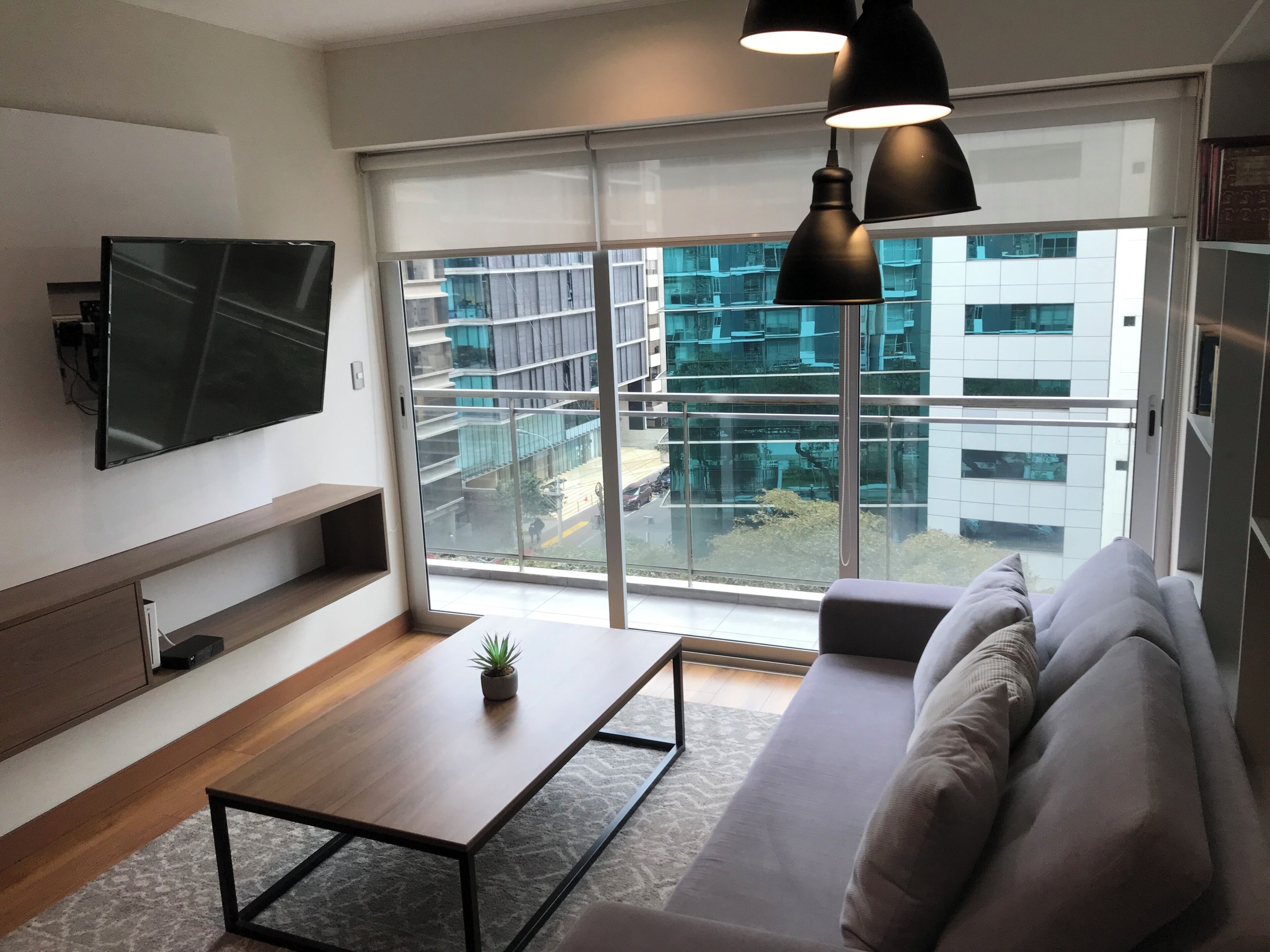 Un soggiorno moderno con una TV a schermo piatto, un divano, un tavolino da caffè e un'ampia finestra che si affaccia su un edificio.