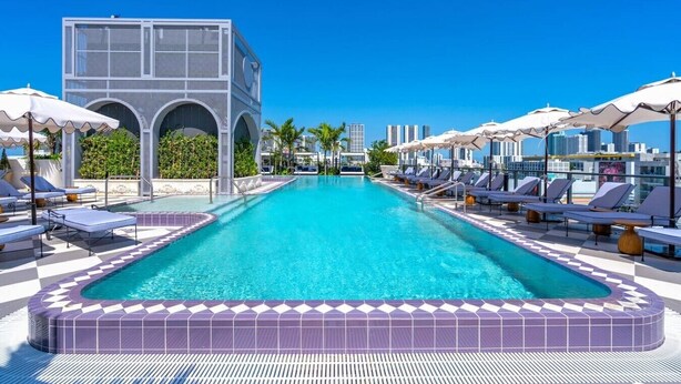 Ein Pool mit Liegestühlen und Sonnenschirmen, ein Gebäude mit Rundbogenfenstern und ein klarer, blauer Himmel.
