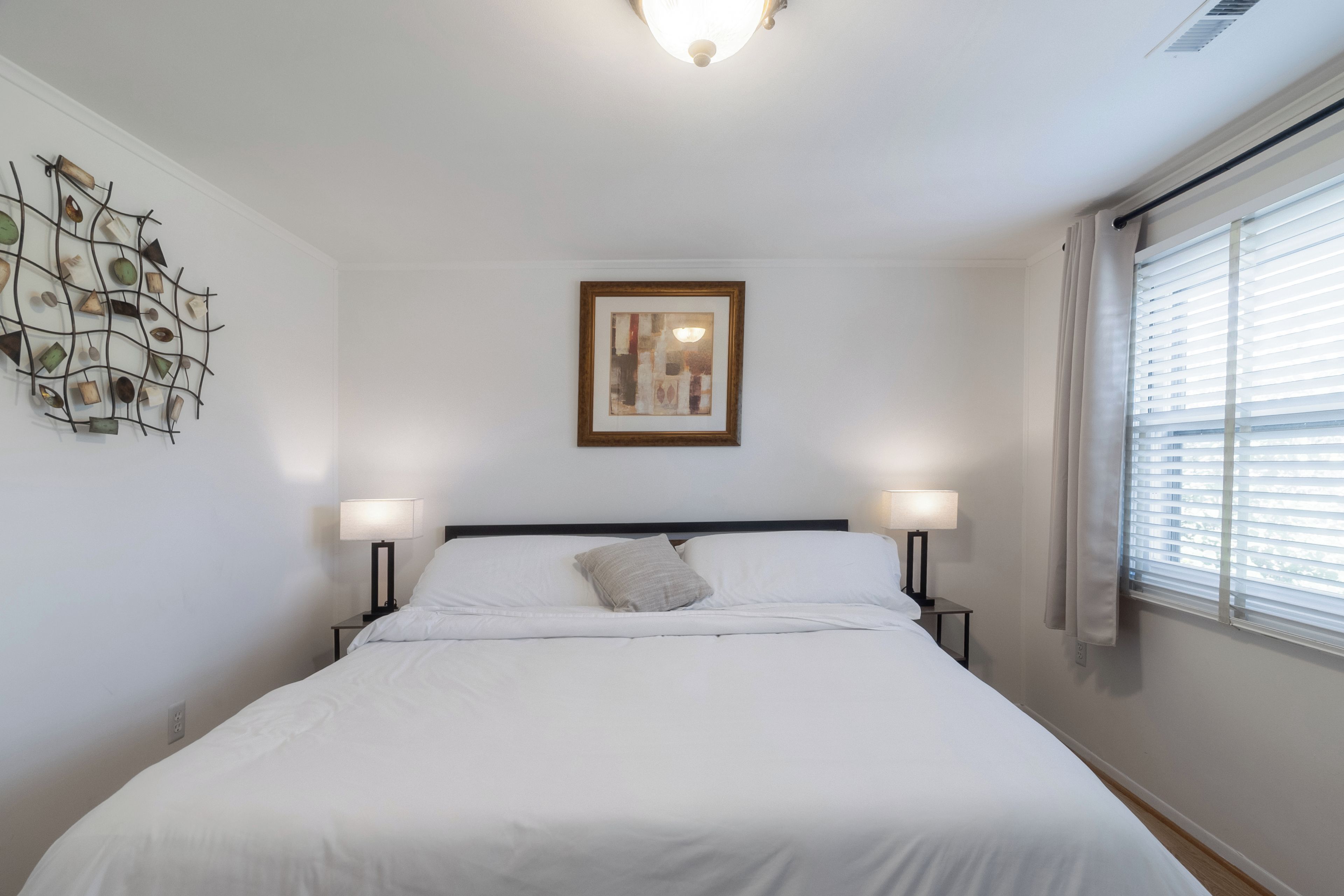A hotel room with a bed, two bedside lamps, a framed picture on the wall, and a window with blinds.
