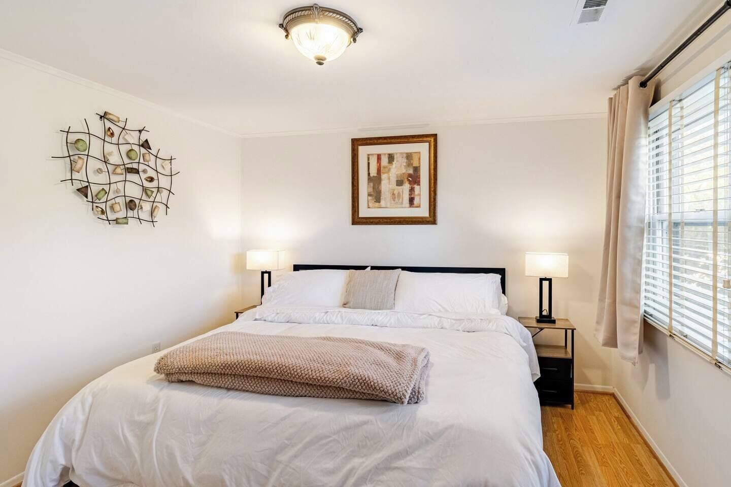 Un dormitorio ordenado con cama, dos lámparas de noche, una obra de arte en la pared y un ventanal con persianas.
