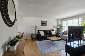 Ein Wohnzimmer mit einer Couch, Couchtisch und Fernseher. Es gibt Pflanzen, einen Spiegel und ein Bild an der Wand.