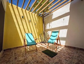 Eine Terrasse mit zwei Holzstühlen, einer grünen Matte und einer gelben Wand mit vertikalen Holzlamellen.