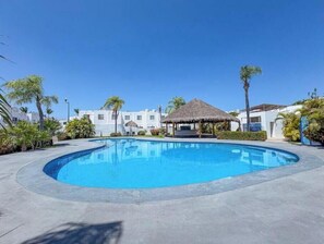 Una piscina con una struttura a tetto di paglia, circondata da palme e edifici bianchi.