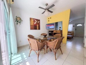 Ein Essbereich mit Korbstühlen, einem Glastisch, einem Deckenventilator, einem Fernseher und einer gelben Akzentwand.