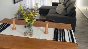 A modern living room with a sofa, a dining table with chairs, a TV mounted on the wall, and a window with sheer curtains.
