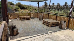 An outdoor patio with wooden benches and a tiled floor, surrounded by a wooden fence and palm trees in the background.