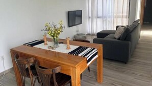 A dining area with a wooden table, chairs, a vase with flowers, a flat-screen TV, and a grey sofa in a modern living space.