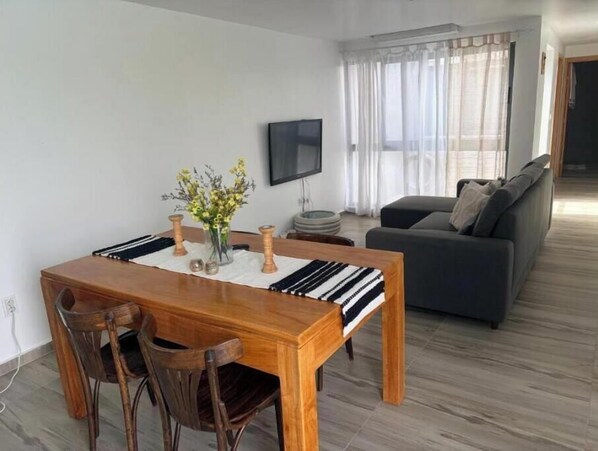 A dining area with a wooden table, chairs, a vase with flowers, a flat-screen TV, and a grey sofa in a modern living space.