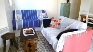 Een woonkamer met een gestreept bankje, een klein houten tafeltje, een kamerkleed met een patroon, een bed met een roze dekbed en een wandklok.