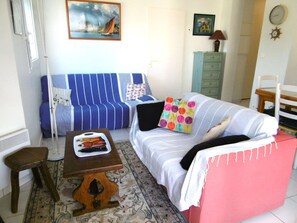 Ein Wohnzimmer mit einem gestreiften Sofa, einem kleinen Holztisch, einem gemusterten Teppich, einem Bett mit rosa Bettwäsche und einer Wanduhr.