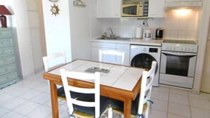 Een keuken met witte kastjes, een magnetron, een koffiezetapparaat, een vaatwasser en een oven. Er staat een eettafel met stoelen.