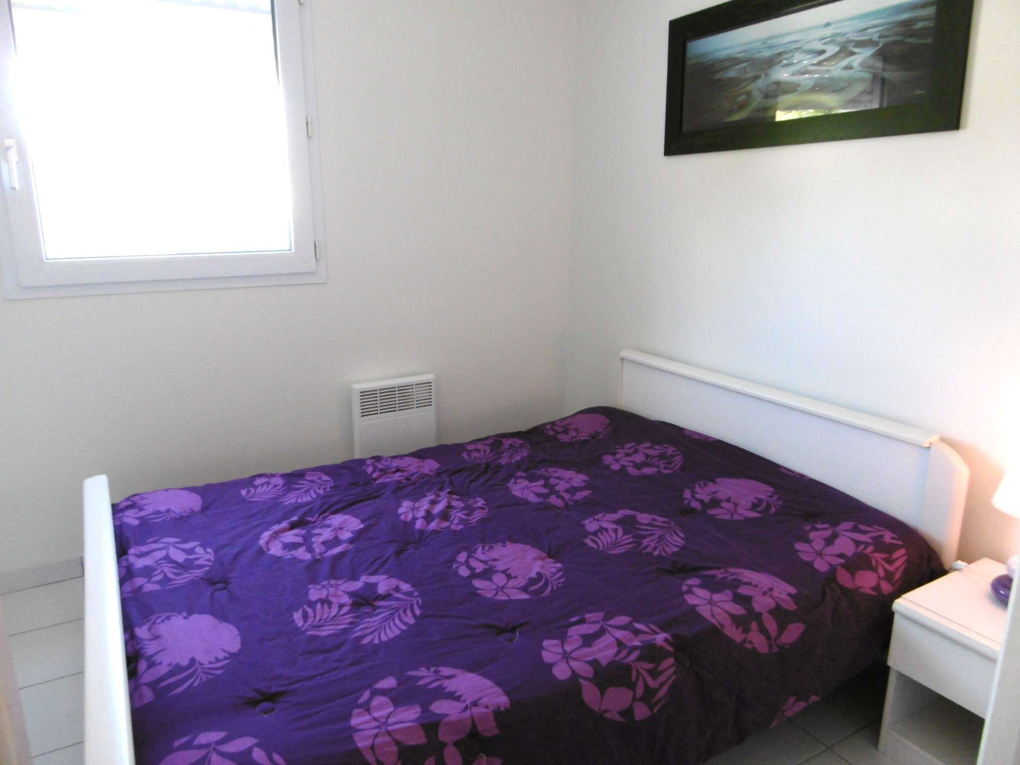 A single bed with a purple floral bedspread, a white bedside table, and a framed picture on the wall.