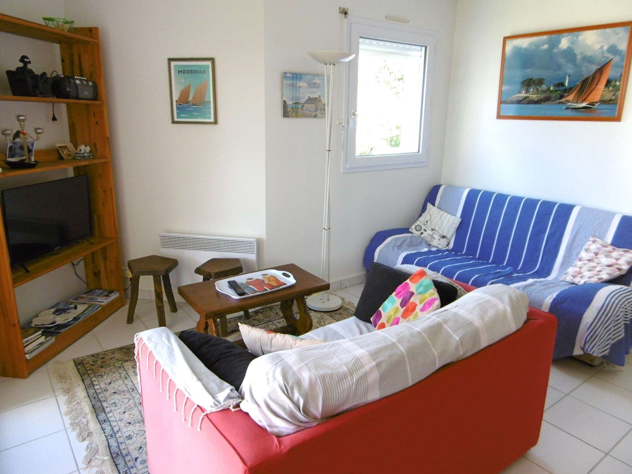 Una sala de estar con un sofá rojo, un sofá azul a rayas, una estantería de madera, una mesa con una bandeja y una ventana con persianas.