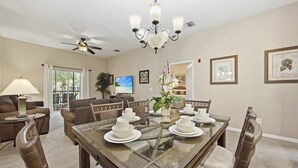 A dining area with a glass-top table, chairs, a chandelier, ceiling fan, and a television in the background. - Family 808 Cozy Poolside Retreat  2BR 2BA (Davenport)