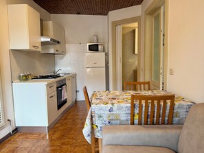 Kompakti keittiö, jossa on liesi, uuni, mikroaaltouuni ja jääkaappi. Viereisessä olohuoneessa on ruokapöytä tuoleineen ja sohva.