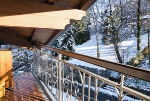 Un balcón de madera con barandilla de metal que da a un paisaje nevado con árboles y un edificio al fondo.