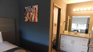 Ein Hotelzimmer mit Bett, einem Wandbild, einem Badezimmer mit Spiegel und einer Küchenzeile mit Schränken und Arbeitsplatte.