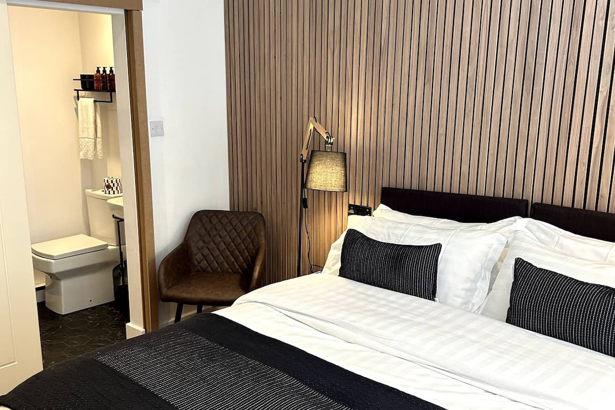 A hotel room with a large bed, a bedside table, a chair, a lamp, and a view of the bathroom.