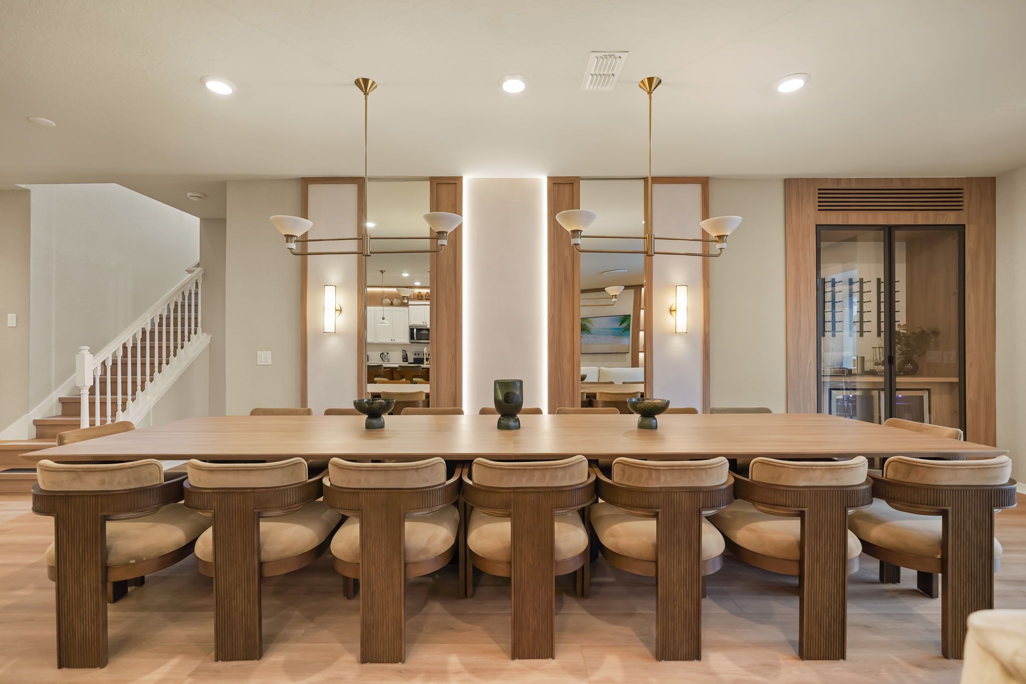 A spacious dining area with a long wooden table and matching chairs, a staircase on the left, and a kitchen visible through a glass door.