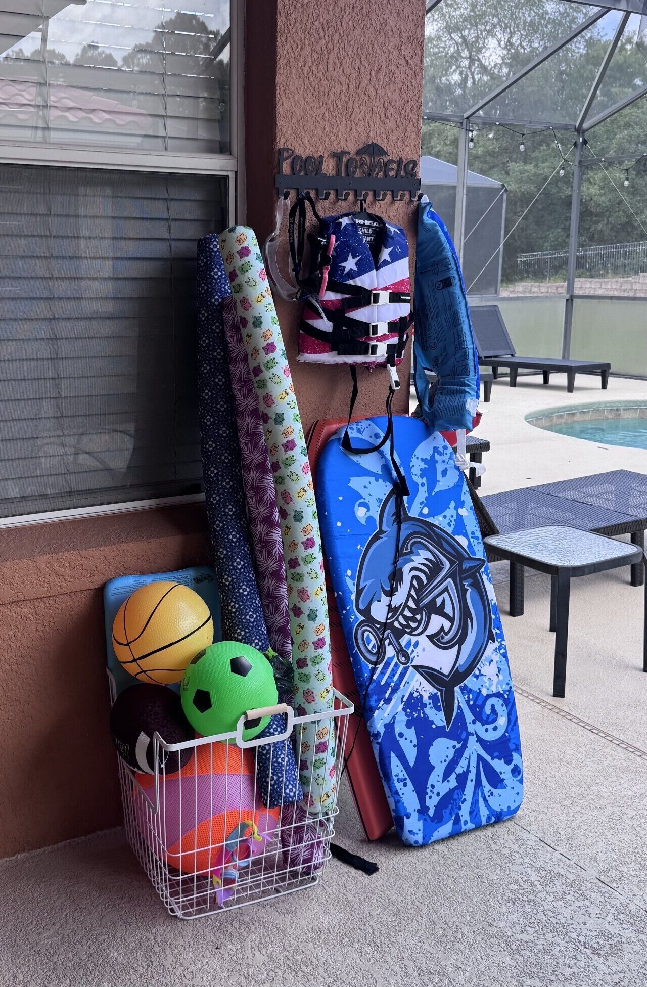 A poolside area with a pool table, lounge chairs, and a metal rack holding sports balls and a blue bag.