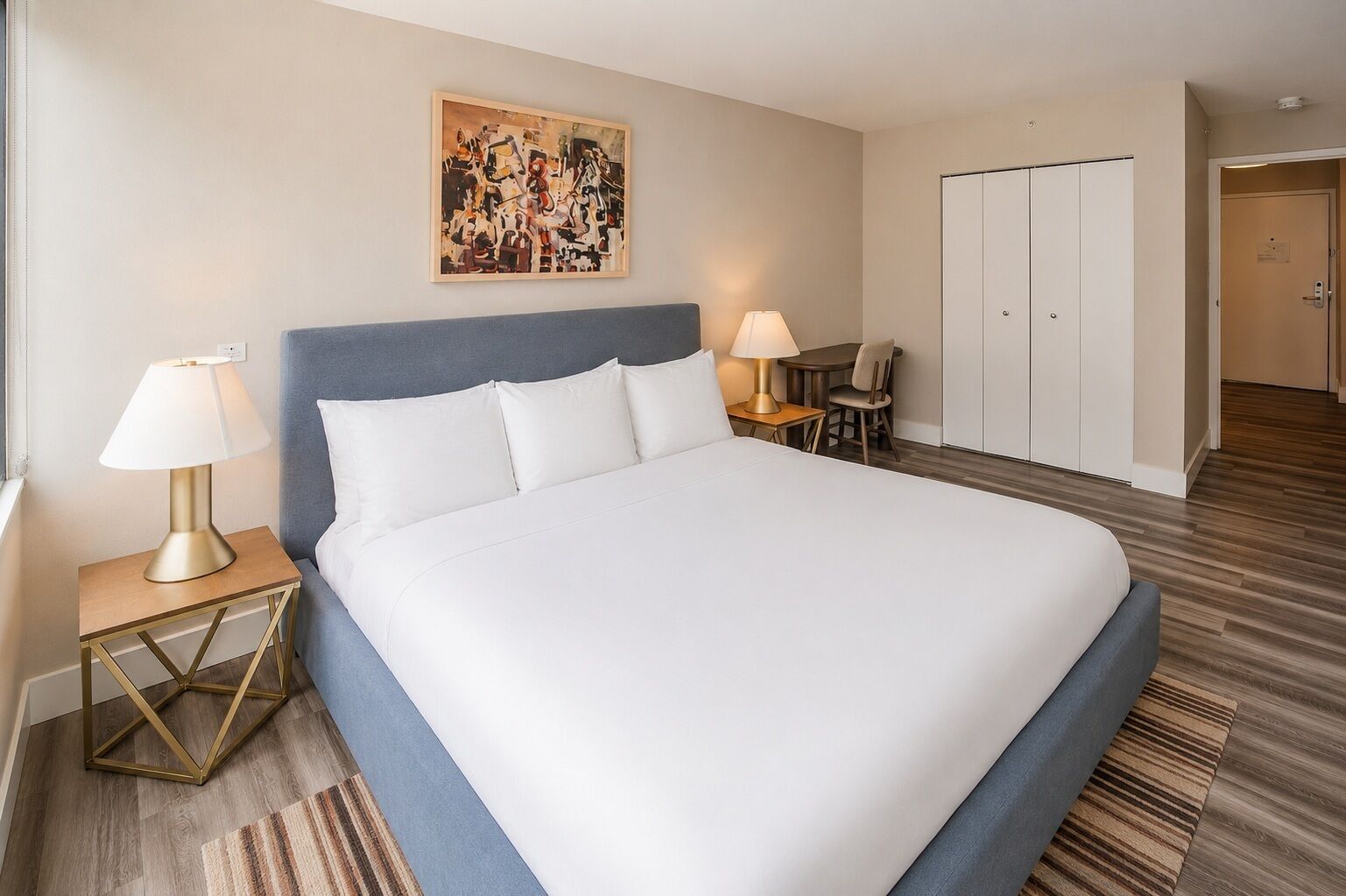 Ein Hotelzimmer mit einem großen Bett, zwei Nachttischen mit Lampen, einem kleinen Tisch mit einem Stuhl, einem Kleiderschrank und einem Bild an der Wand.