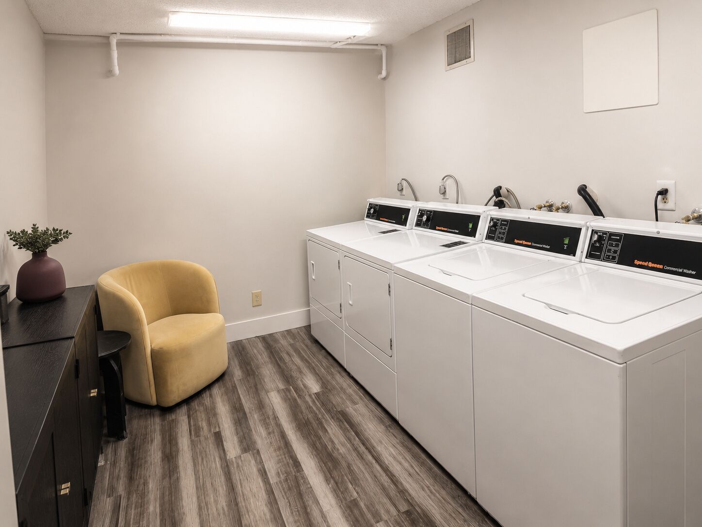 A laundry room with three white commercial washers and dryers, a yellow chair, and a potted plant.