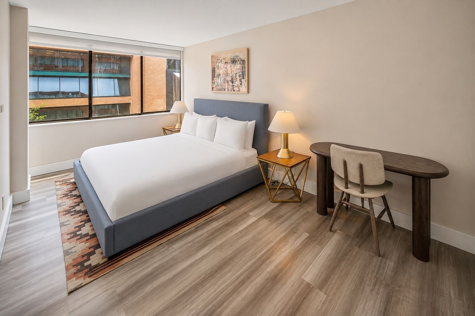 A hotel room with a bed, a desk with a chair, a lamp, and a window with a view of buildings.