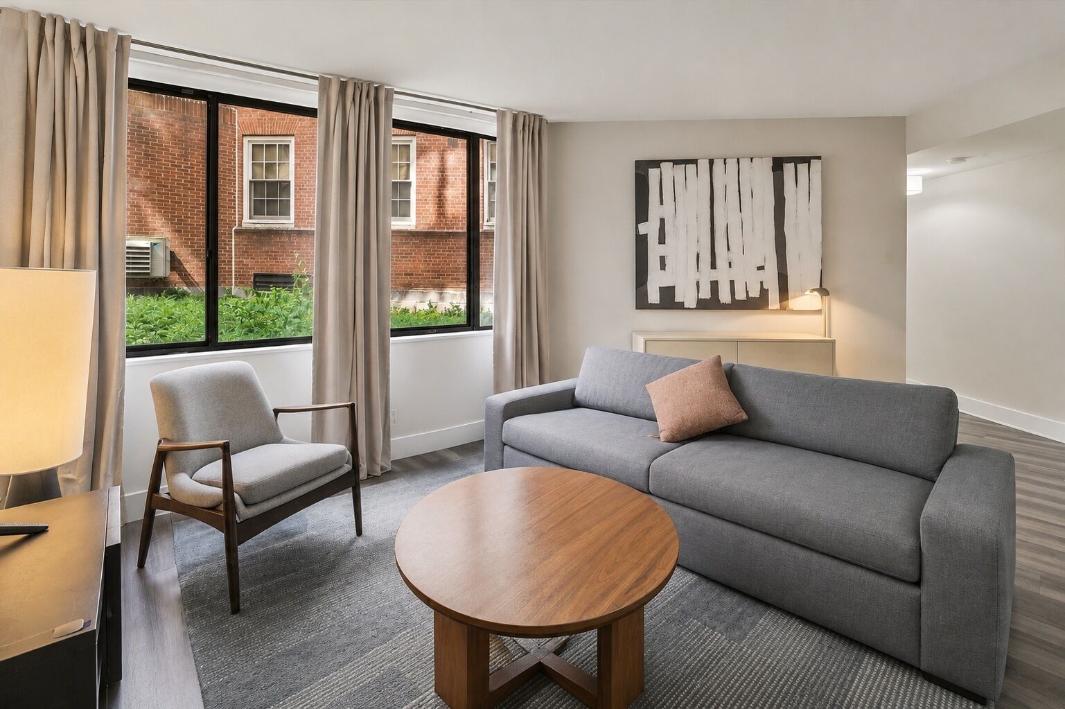 Quarto de hotel com um sofá cinzento, uma mesa de centro de madeira, uma cadeira e uma janela com cortinas.