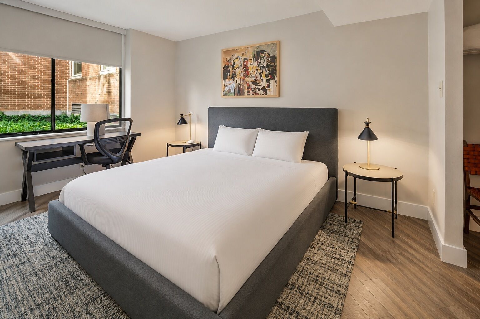 Quarto de hotel com uma cama grande, uma escrivaninha, duas cadeiras, uma janela com vista para o verde e um quadro na parede.