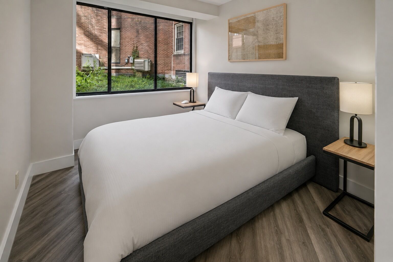 Une chambre d’hôtel avec un grand lit, une table de chevet avec une lampe, une fenêtre donnant sur un immeuble en briques et un tableau accroché au mur.