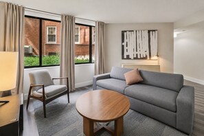 Ein Hotelzimmer mit grauem Sofa, einem Holztisch, einem Sessel und einem Fenster mit Vorhängen.