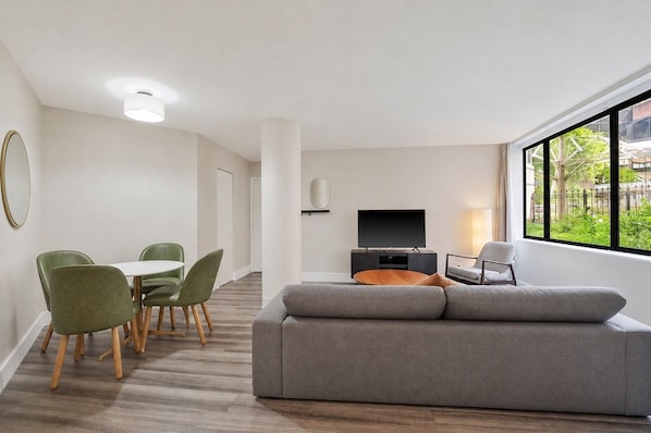 Ein modernes Wohnzimmer mit einem grauen Sofa, einem Fernseher und einem Essbereich mit grünen Stühlen.