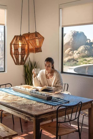 Eine Person liest an einem Tisch mit großem Fenster, einer gewebten Pendelleuchte und Blick auf Felsen.