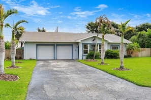 A single-story house with a garage, a front lawn with palm trees, and a clear blue sky. - Tropical Backyard Oasis in Cocoa Beach | Heated Pool, BBQ Grill & Fire Pit ☀️ (Cocoa Beach)