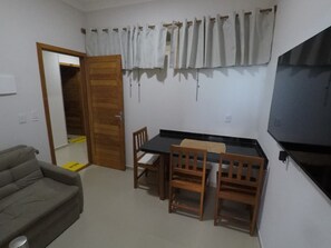 Una habitación pequeña con una mesa de comedor de madera y sillas, un sofá gris, un televisor montado en la pared y una puerta que da a otra habitación.