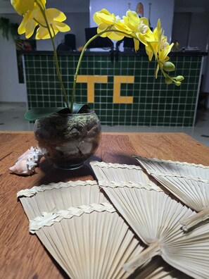 A vase with yellow flowers on a wooden table, a tiled wall with a letter 'TE' in the background, and a decorative fan. - TC FLATS (Itacaré)