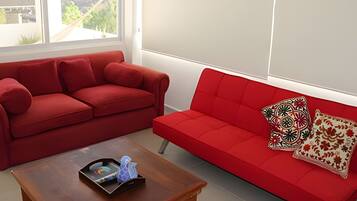 A red sofa with patterned pillows, a wooden coffee table with a tray and decorative items, and a view of greenery through a window.