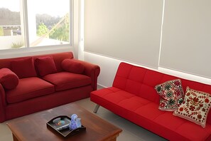 A red sofa with patterned pillows, a wooden coffee table with a tray and decorative items, and a view of greenery through a window. - BUDA Playa Salchi Valentina (San Pedro Pochutla)