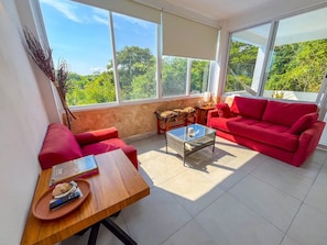A sunlit room with a red sofa, a wooden coffee table, and a view of greenery outside.