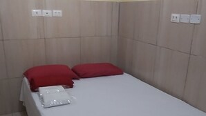 A single bed with a white bedspread and red pillows, a small rug, and a wall with electrical outlets.